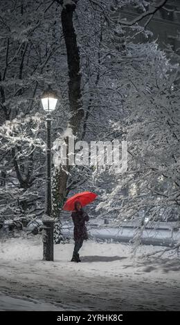 Eine Person steht in einer verschneiten Landschaft in Genf, Schweiz, mit einem leuchtenden roten Regenschirm unter einer leuchtenden Straßenlaterne, schneebedeckte Bäume und Boden Stockfoto