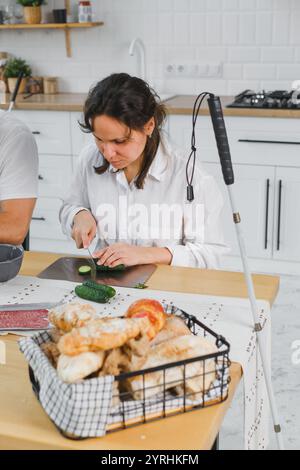 Eine junge blinde Frau hackt Gurken in einer hellen, modernen Küche. Ein Brotlaib ist in einem Korb in der Nähe. Ein Stock deutet auf Sehstörungen hin. Inklusive c Stockfoto