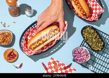 Eine Nahaufnahme von Händen, die einen Hotdog mit Senf halten, umgeben von bunten Belägen auf einem hellblauen Hintergrund Stockfoto