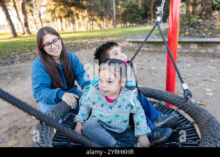 Eine Familie verbringt schöne Zeit in einem Park die ältere Schwester lächelt, während sie ihre Kinder auf einer Schaukel spielen sieht Stockfoto