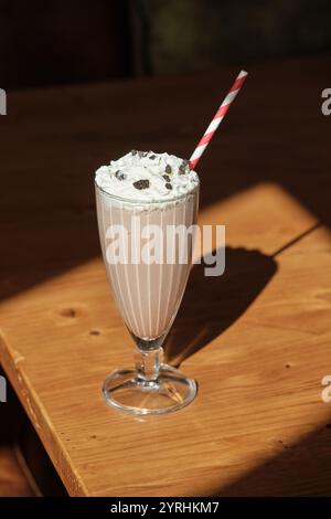 Ein köstlicher Vanillemilkshake mit Schlagsahne und Schokoladenchips wird elegant in einem Vintage-Glas serviert. Ein weiß-rot gestreifter Strohhalm sorgt für einen Spielspaß Stockfoto