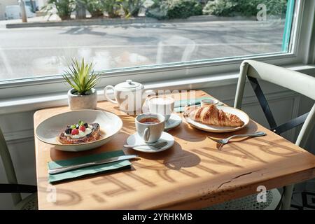 Der sonnendurchflutete Café-Tisch bietet köstliche Snacks, darunter ein Croissant, französischer Toast mit Beeren, eine Teekanne und zwei Tassen Kaffee, Die gemütlich und einladend Sind Stockfoto