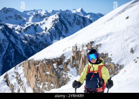 Eine Skifahrerin mit Helm und Spiegelbrille steht auf einem schneebedeckten Berghang und lächelt vor der Kulisse der schroffen Gipfel unter einem klaren blauen Sch Stockfoto