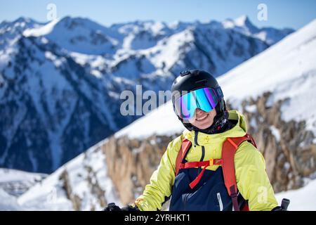 Eine Frau in einem hellen Ski-Outfit steht mit Skistöcken und lächelt vor der Kulisse atemberaubender verschneiten Berge, klarer Himmel und lebendige Ausrüstung Stockfoto