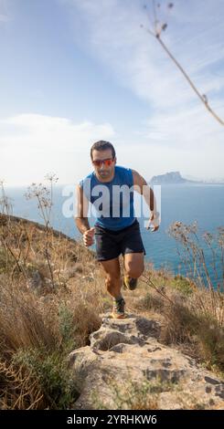 Ein Mann in sportlicher Kleidung läuft auf einem felsigen Pfad entlang der Küste. Das pulsierende Meer und die fernen Berge bilden eine atemberaubende Kulisse, die den Geist von fängt Stockfoto