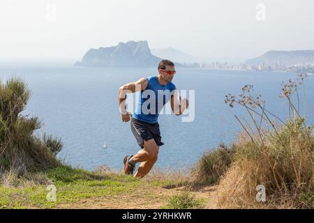 Ein Mann in sportlicher Kleidung läuft auf einem malerischen Küstenweg. Der Hintergrund bietet einen atemberaubenden blick auf das Meer und die fernen Berge unter einem klaren Himmel Stockfoto