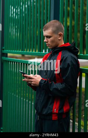 Ein junger Mann in einer schwarz-roten Sportjacke steht an einem grünen Metallzaun und konzentriert sich auf sein Smartphone. Das Ambiente im Freien ist lässig und hebt den modernen Charakter hervor Stockfoto