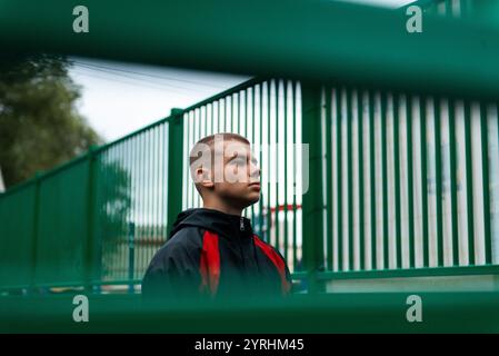 Ein junger Mann in einer schwarz-roten Jacke steht im Profil vor einem grünen Metallzaun und vermittelt ein Gefühl der Besinnung und des städtischen Lebens. Der Fokus liegt auf o Stockfoto