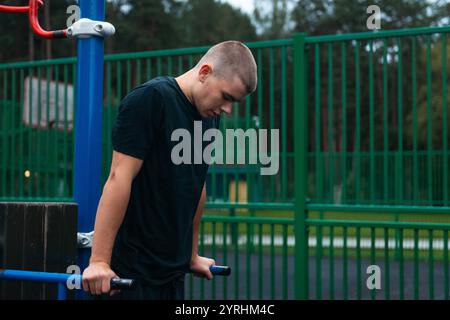 Ein junger Mann in einem schwarzen T-Shirt übt an parallelen Bars in einem Park im Freien. Der grüne Metallzaun und die Bäume im Hintergrund schaffen eine ruhige Atmosphäre Stockfoto