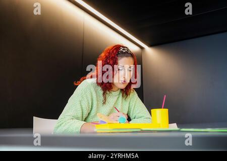 Eine Transfrau mit roten Haaren arbeitet intensiv an einem Schreibtisch, umgeben von Kunstgegenständen, in einem modernen Arbeitsbereich. Die Umgebung unterstreicht Kreativität und Vielfalt Stockfoto