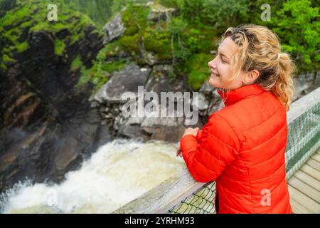 Eine Frau in einer knallroten Jacke genießt die üppige, malerische Aussicht auf Schwedisch Lappland, die auf einer Holzbrücke mit Blick auf einen rauschenden Wasserfall im wi steht Stockfoto