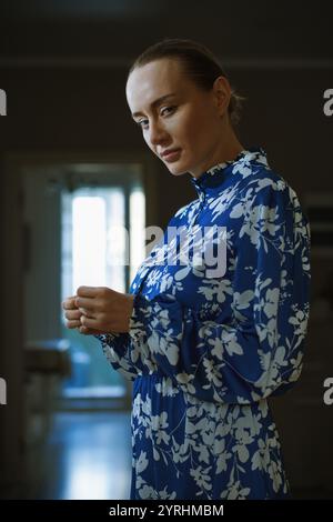 Eine Frau in einem blauen Blumenkleid steht neben einem sonnendurchfluteten Fenster. Das sanfte Leuchten hebt ihren kontemplativen Ausdruck hervor und schafft eine heitere und intime Atmosphäre Stockfoto
