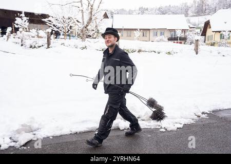 Ein fröhlicher Schornsteinfeger geht selbstbewusst durch eine schneebedeckte Dorfstraße und trägt seine Putzwerkzeuge. Die heitere Winterszene zeigt ein Q Stockfoto