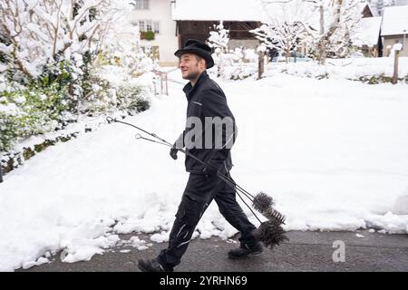 Ein fröhlicher Schornsteinfeger geht selbstbewusst durch eine schneebedeckte Dorfstraße und trägt seine Putzwerkzeuge. Die heitere Winterszene zeigt ein Q Stockfoto