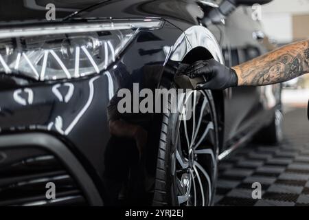 Der Mann wischt die Reifen eines schwarzen Autos mit einem Schwamm in einer Garage ab. Waschen und Pflege der Räder. Gummipolitur Stockfoto
