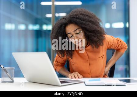 Junge Geschäftsfrau, die sich aufgrund von Rückenschmerzen am Schreibtisch unwohl fühlt. Sie lehnt sich bei der Arbeit an einem Laptop nach vorne und hebt die Herausforderungen an die Ergonomie am Arbeitsplatz hervor. Stress und Unwohlsein sichtbar. Stockfoto