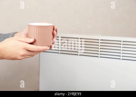 Die Frau friert zu Hause. Er sitzt neben einem elektrischen Heizkörper mit einer Tasse heißem Tee in den Händen. Stockfoto