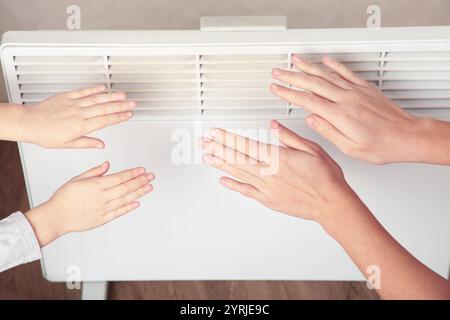 Eine junge Frau und ein kleines Mädchen, die Hände in der Nähe einer modernen elektrischen Heizung wärmen. Draufsicht Stockfoto