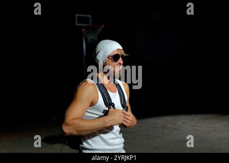 Eine muskulöse Person posiert bei schwachem Licht, trägt eine Sonnenbrille und eine Mütze, während sie den Rucksack hält. Seine selbstbewusste Haltung spiegelt einen mächtigen und styl wider Stockfoto