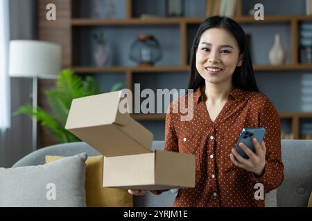 Glückliche asiatische Frau, die zu Hause Telefon und Kartonschachteln hält und moderne Technologie und Bequemlichkeit demonstriert. Erfasst das Wesen von E-Commerce und Online-Shopping. Stockfoto