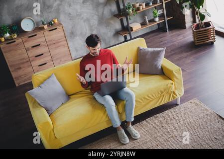 Junger Mann, der ein entspanntes Wochenende genießt, auf einem gelben Sofa sitzt, an einem Laptop in einer komfortablen Wohnung mit Tageslicht arbeitet. Stockfoto