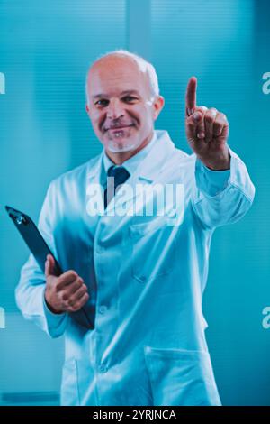 Ein reifer männlicher Arzt, der einen Labormantel trägt, hält ein Klemmbrett und zeigt seinen Zeigefinger nach oben, was eine wichtige medizinische Anweisung oder Aufnahme bedeutet Stockfoto