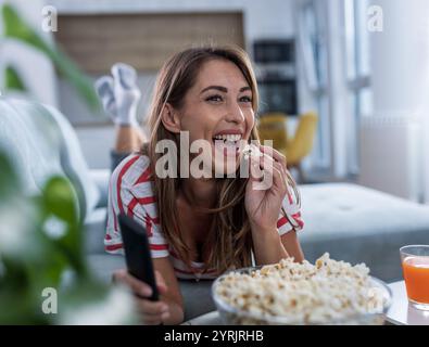 Lächelnde junge Frau, die Fernbedienung hält, Filme ansieht und Popcorn zu Hause im gemütlichen Wohnzimmer isst Stockfoto