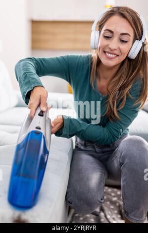 Glückliche junge Frau, die einen Handstaubsauger hält und Staub auf dem Sofa im Wohnzimmer feststeckt, während sie Musik über das Headset hört Stockfoto