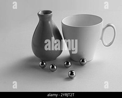Ein auffälliges monochromes Stillleben-Foto mit einer weißen Keramikschale, einer glatten Vase und reflektierenden Metallkugeln, die künstlerisch angeordnet sind. Stockfoto