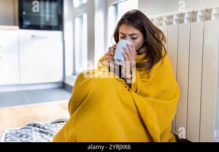 Hübsches Mädchen, das friert, auf dem Boden sitzt, in Decke gehüllt, sich auf den Heizkörper lehnt und heißen Tee aus der Tasse trinkt Stockfoto