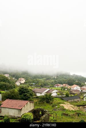 Ein ländliches Dorf mit verstreuten Häusern, umgeben von üppiger grüner Vegetation und Hügeln. Eine nebelige Atmosphäre bedeckt die Landschaft und schafft ein ruhiges und angenehmes ambiente Stockfoto