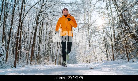 Lächelnder Trailläufer mittlerer Alters, gekleidet in leuchtend orangefarbener winddichter Jacke, hält an einem sonnigen, frostigen Tag schnell durch malerische, verschneite Wälder. S Stockfoto