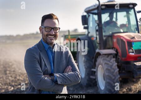 Porträt eines lächelnden, selbstbewussten Geschäftsmannes mit gekreuzten Armen vor dem Traktor im Herbst auf dem Feld. Bankvertreter und Kreditkonzept für kleine Unternehmen Stockfoto