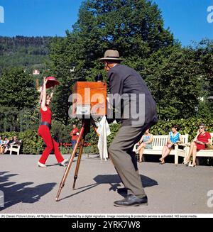 Fotograf und Model der 1960er Jahre. Die Schauspielerin und Sängerin Grynet Molvig fotografierte in der norwegischen Stadt Bergen und posierte für einen Fotografen mit seiner antiken Kamera auf einem Stativ. Grynet ist ihr rotes Outfit, hebt ihren Hut und lächelt. 1969 Stockfoto