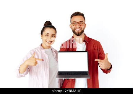 Positiver Mann und Frau arabischer oder kaukasischer Abstammung, der einen Laptop mit einem leeren weißen Bildschirm hält, darauf zeigt, Inhalte oder Produkte fördert, auf isoliertem weißen Hintergrund steht und lächelt Stockfoto