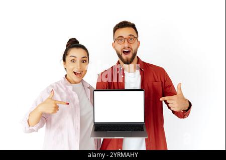 Aufgeregter Mann und Frau arabischer oder kaukasischer Herkunft, die einen Laptop mit leerem Modellbildschirm halten, lächeln und darauf zeigen, Inhalte oder Produkte bewerben, stehen auf einem isolierten weißen Hintergrund Stockfoto