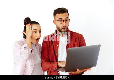 Aufgeregter Mann und Frau, arabische oder kaukasische Ethnie, beide starren auf einen Laptop-Bildschirm mit weit geöffneten Augen und schockiertem Ausdruck, lässig gekleidet, stehend auf isoliertem weißen Hintergrund Stockfoto