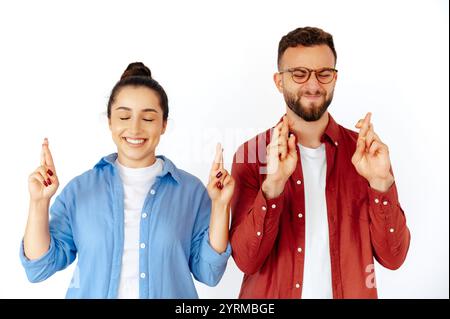 Hoffnungsvolle Mann und Frau, die mit geschlossenen Augen die Finger kreuzen und viel Glück wünschen, gekleidet in lässigen roten und blauen Hemden, stehen auf isoliertem weißem Hintergrund Stockfoto