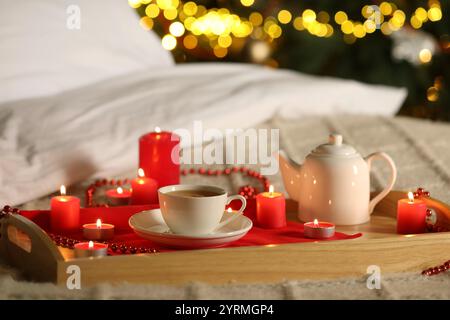 Aromatischer Tee in der Tasse, Teekanne und brennende Kerzen auf dem Bett gegen verschwommenes Weihnachtslicht, Bokeh-Effekt Stockfoto