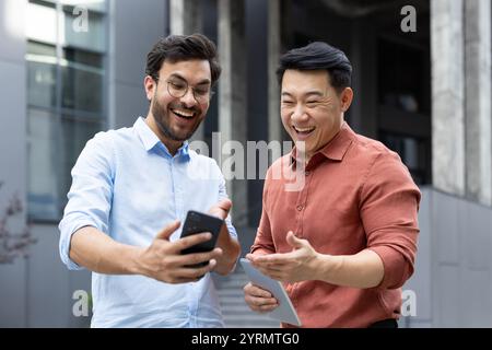 Zwei Kollegen teilen einen freudigen Moment draußen und interagieren mit einem Smartphone und Tablet. Die Szene fängt eine freundliche, energiegeladene Atmosphäre ein, die moderne Technologie und Kommunikation hervorhebt. Stockfoto