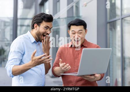 Zwei ekstatische Kollegen feiern einen überraschenden Erfolg, während sie sich einen Laptop-Bildschirm ansehen. Ihr fröhlicher Ausdruck und das Notebook zeigen einen Moment der Leistung und Teamarbeit. Stockfoto