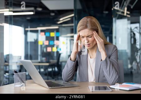 Eine reife Geschäftsfrau fühlt sich gestresst, wenn sie in einem modernen Büro arbeitet. Sie sitzt an einem Schreibtisch mit Laptop und Telefon. Ihre Hände drücken gegen ihre Schläfe und vermitteln Spannung. Stockfoto