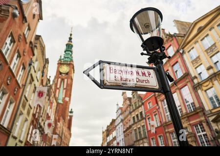 Ein Straßenschild, das auf die Marienkirche in Gdańsk (Polen) zeigt, mit einem historischen Uhrenturm im Hintergrund und farbenfrohen Gebäuden entlang der Straße. Stockfoto