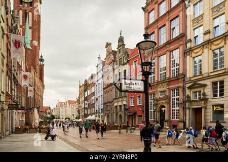 Ein Straßenschild, das auf die Marienkirche in Gdańsk (Polen) zeigt, mit einem historischen Uhrenturm im Hintergrund und farbenfrohen Gebäuden entlang der Straße. Stockfoto
