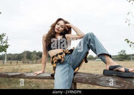 Schöne, frische herbstliche Landfrau in stilvollem kariertem Hemd mit Vintage Blue Jeans sitzt und posiert auf einem Holzzaun auf dem Land Stockfoto