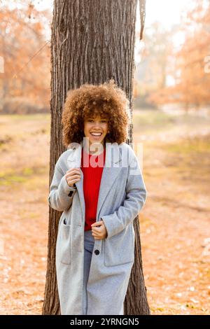 Eine junge Frau mit lockigen Haaren lehnt sich an einen Baum, trägt einen warmen Pullover, lächelt fröhlich im goldenen Herbstlaub und umhergefallenen Blättern Stockfoto