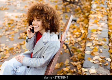 Eine junge Frau mit lockigen Haaren lacht fröhlich, während sie auf einer Parkbank sitzt, die mit lebhaften Herbstblättern bedeckt ist Stockfoto