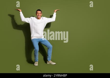 Fröhlicher junger Mann in einem stylischen weißen Pullover posiert verspielt vor khakifarbenem Hintergrund, Arme hoch in einer lustigen Geste Stockfoto