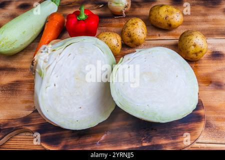 Weißkohl, in zwei Hälften oder Stücke geschnitten, auf einem Holztisch mit Kartoffeln, Paprika, Karotten und Zucchini. Hochwertige Fotos Stockfoto