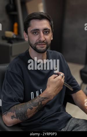 Mann Friseur-Profi sitzt im Stuhl und hält Rasierer zum Rasieren im Friseur Stockfoto
